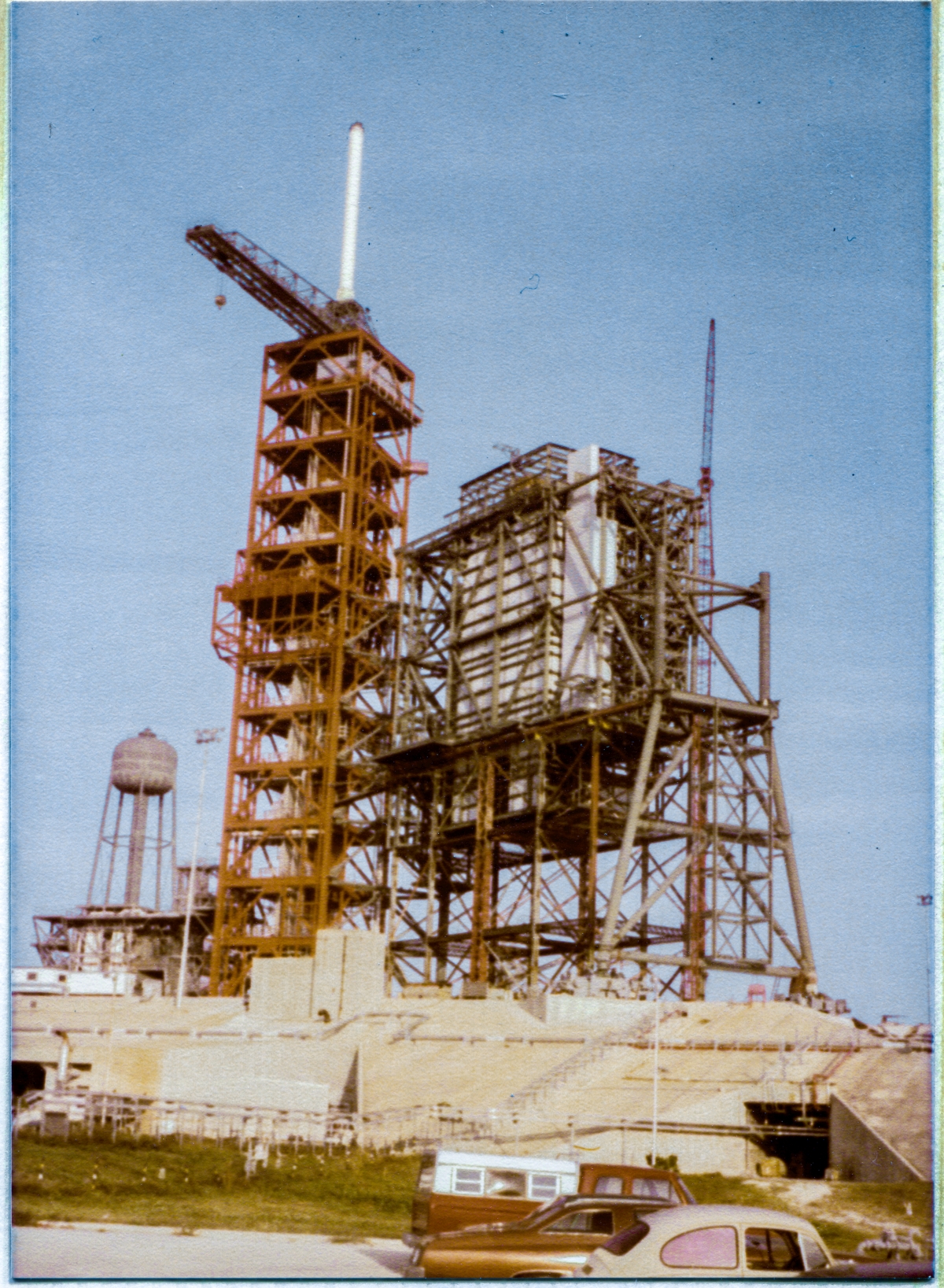 Image 006. Viewed from in front of Sheffield Steel’s field trailer, the structures on Space Shuttle Launch Complex 39-B, Kennedy Space Center, Florida, can be seen, squarely front-lit in the late afternoon. Visible in this image, you can see the concrete structure of the pad itself, the Sound Suppression System water tower, the red multi-platformed structure of the FSS, and the growing steel framework of the RSS still resting on the falsework which will continue to support it until it becomes sufficiently-complete to carry its own weight, allowing for the removal of the falsework. Photo by James MacLaren.